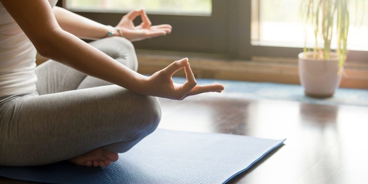 Yoga at home: meditating close-up Attractive young woman working out at home, doing yoga exercise on blue mat, sitting in Easy (Decent, Pleasant Posture), meditating, breathing, relaxing. Body close-up, focus on hand